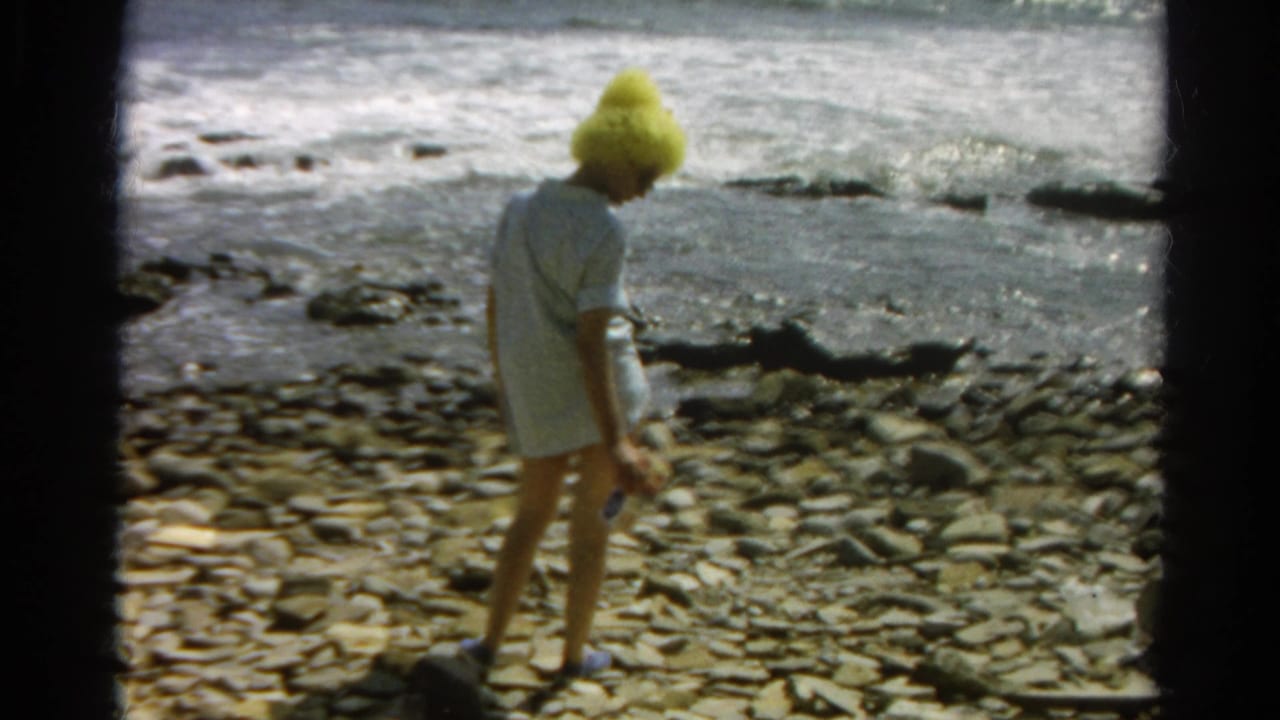 In supported 1966 California home-movie footage, a woman in a yellow hat walks over stones beside surf.