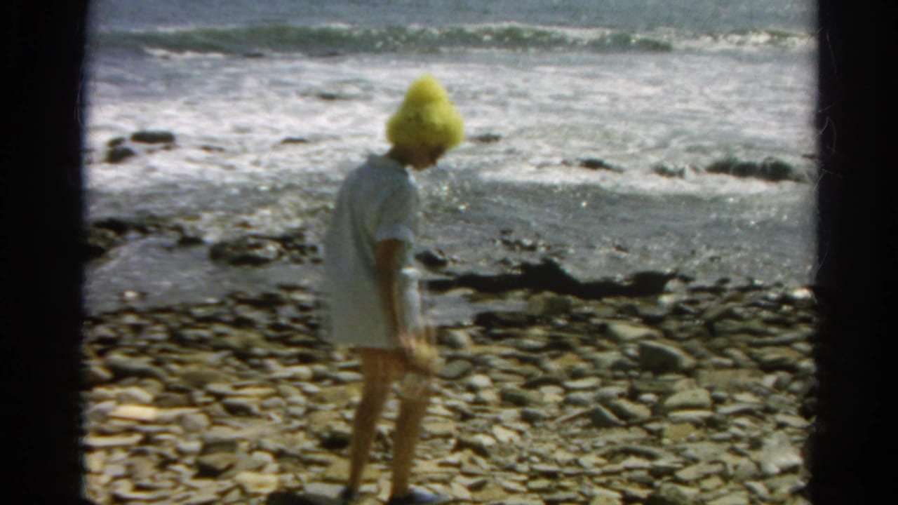 A woman in a yellow hat pauses on a rocky California shoreline, with breaking waves behind her.