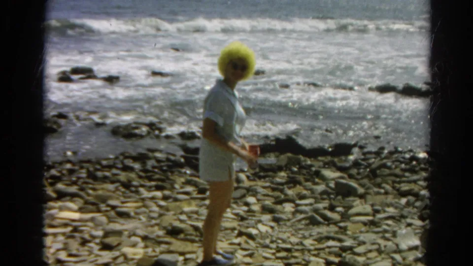 In the 1966 home-movie frame, the woman appears to hold a small object while standing on shoreline stones.