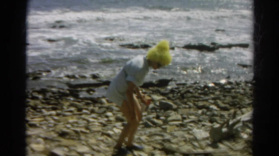 The woman bends down toward stones on the California shoreline, with surf and dark rocks in the background.