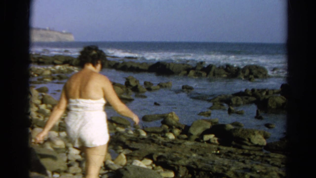 Woman in a white bathing suit steps across rocks beside tide pools and ocean surf.