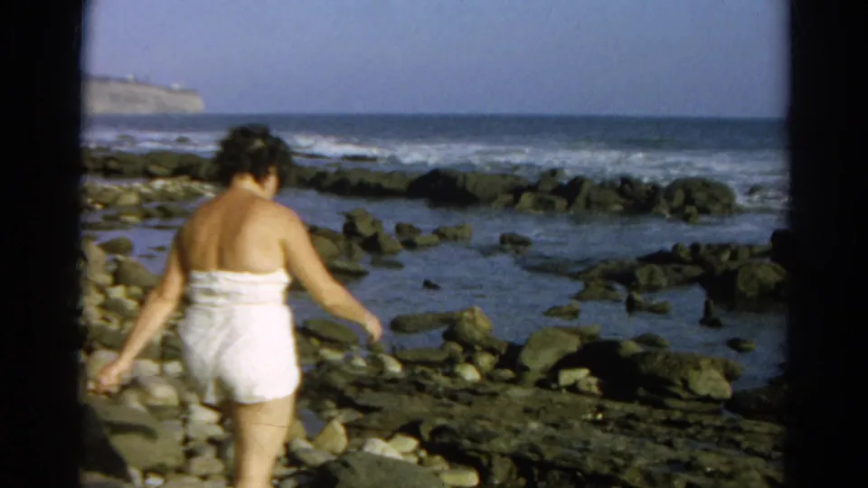 In 1966 home-movie footage identified as Silver Lake, California, a woman in a white bathing suit walks carefully over shoreline rocks near the surf.