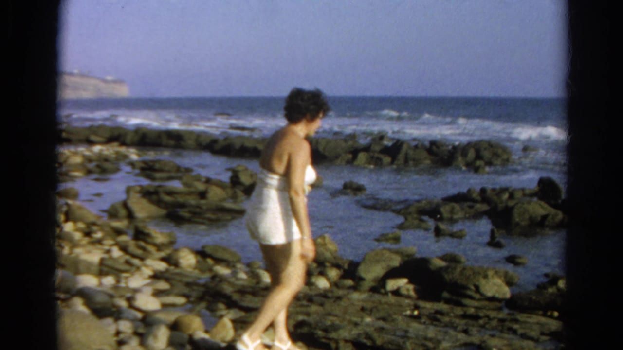 Woman in a white bathing suit walks sideways over dark shoreline rocks near waves.