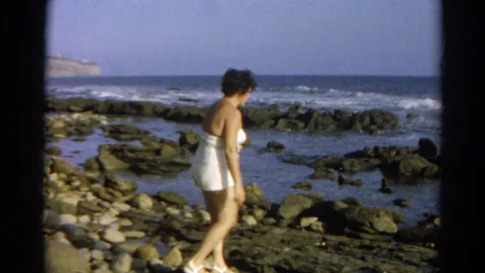 The woman continues across the rocky shore, with shallow pools, breaking waves, and a distant coastal bluff visible in the 1966 clip.