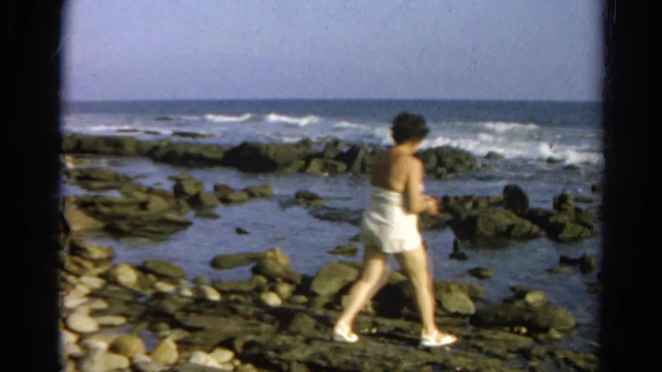 A handheld 8mm frame shows the woman stepping across a flat rock shelf at the shoreline, with tide pools and ocean surf in the background.