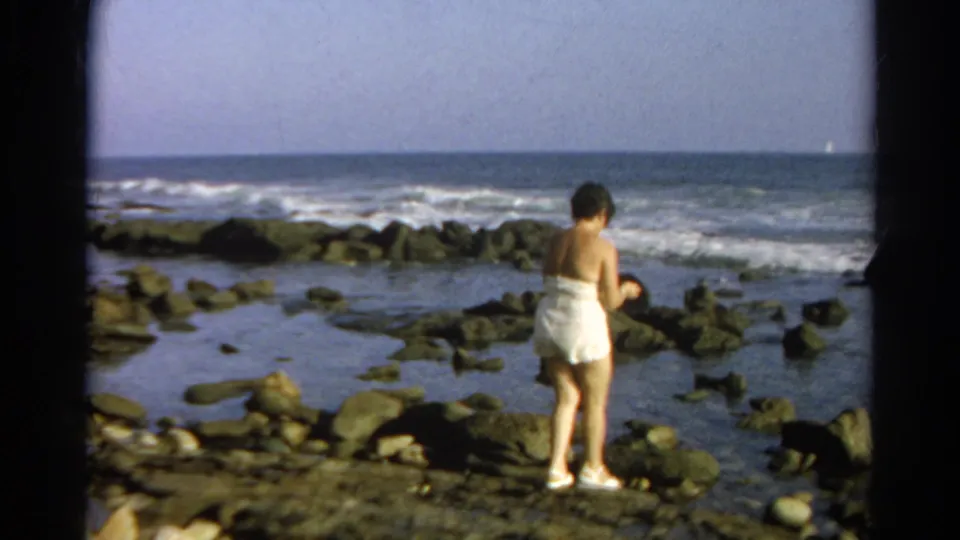 The final frame holds on the woman near the tide pools, facing the water as waves roll across the rocky shoreline.