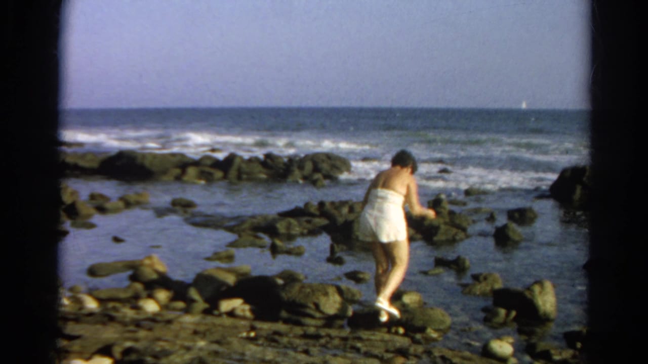 Woman in a white swimsuit steps across rocks beside tide pools and surf.