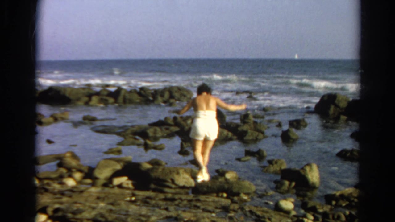 Woman balances on rocks near shallow tide pools with waves behind her.