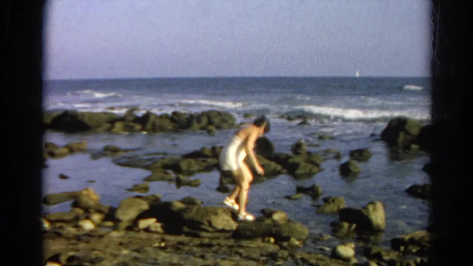 Cataloged 1966 coastal home-movie frame showing a woman bending as she navigates rocks around tide pools near the surf.