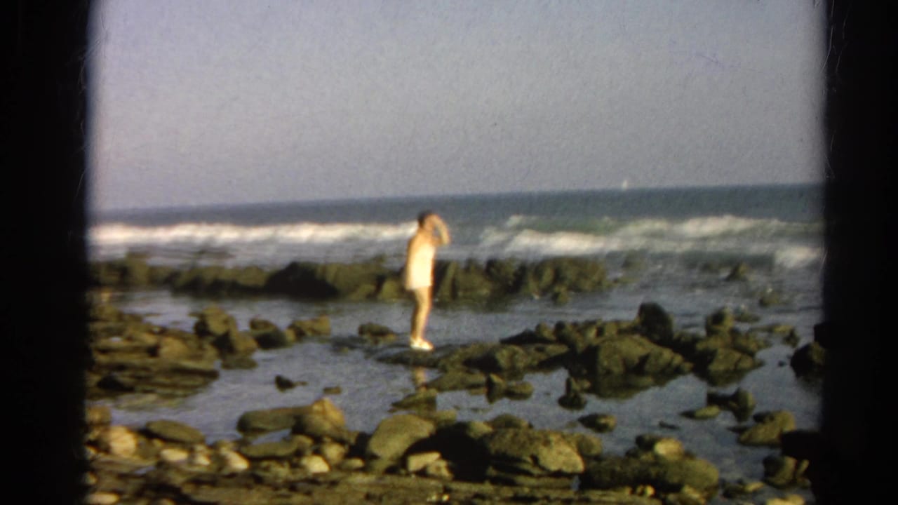Adult woman in a light bathing suit stands on dark rocks near breaking ocean waves.