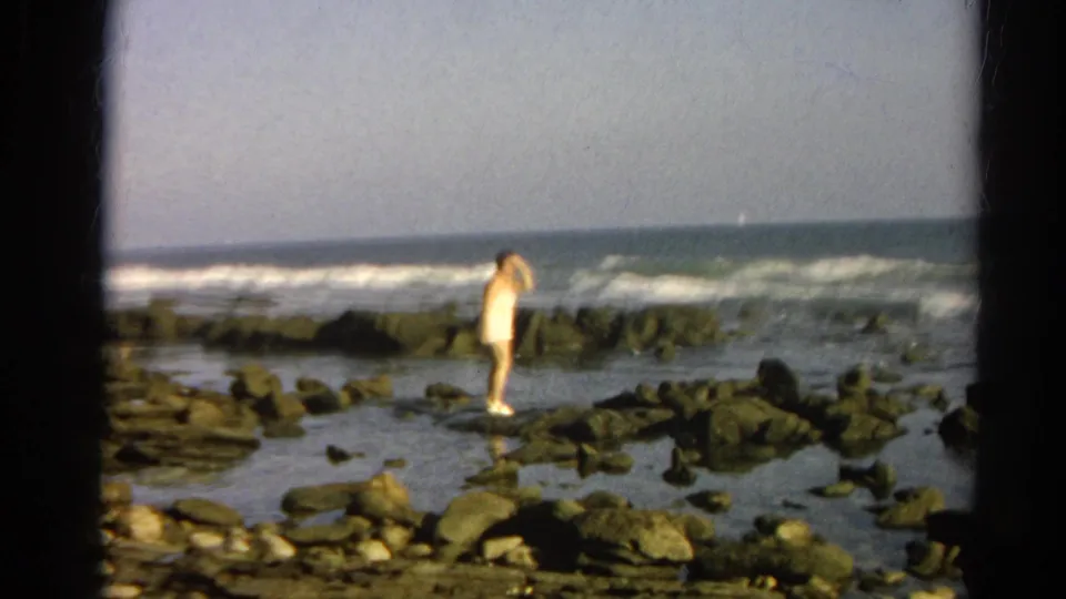 Cataloged 1966 home-movie frame showing an adult woman in a light bathing suit on wet shoreline rocks with surf behind her.