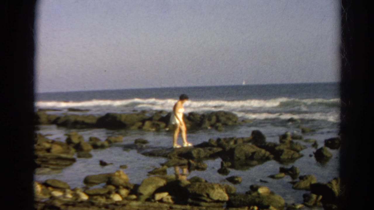 Adult woman in a light bathing suit steps carefully across coastal rocks in cataloged 1966 8mm footage.