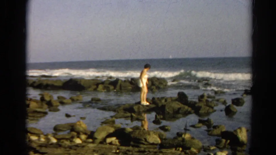 Cataloged 1966 coastal home-movie frame of a woman standing on dark tide-pool rocks near the ocean.