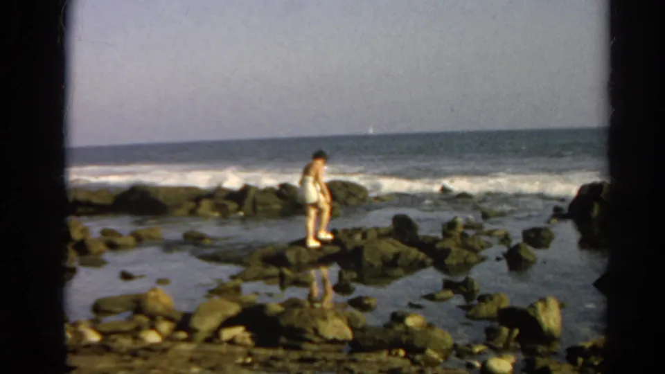 Adult woman in a light bathing suit appears to check her footing on rocky tide pools in 1966 home-movie footage.