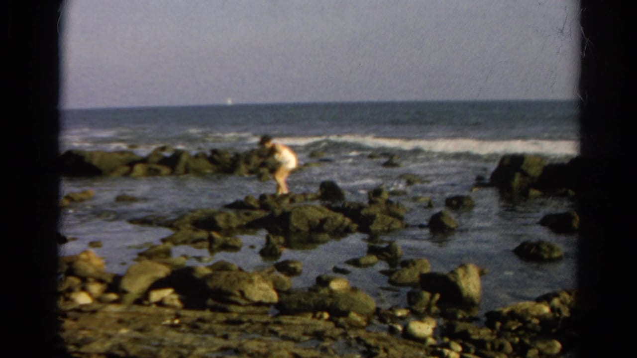 Person in a white swimsuit walking on dark rocks beside ocean surf.