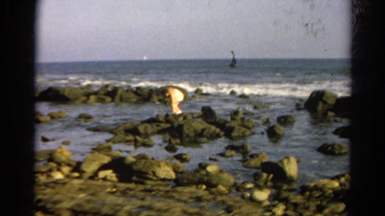 Identified 1966 California 8mm footage shows a person near ocean rocks, partly bent forward as surf breaks behind them.