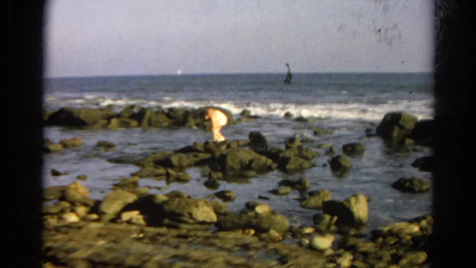 Identified 1966 California 8mm footage shows a person near ocean rocks, partly bent forward as surf breaks behind them.