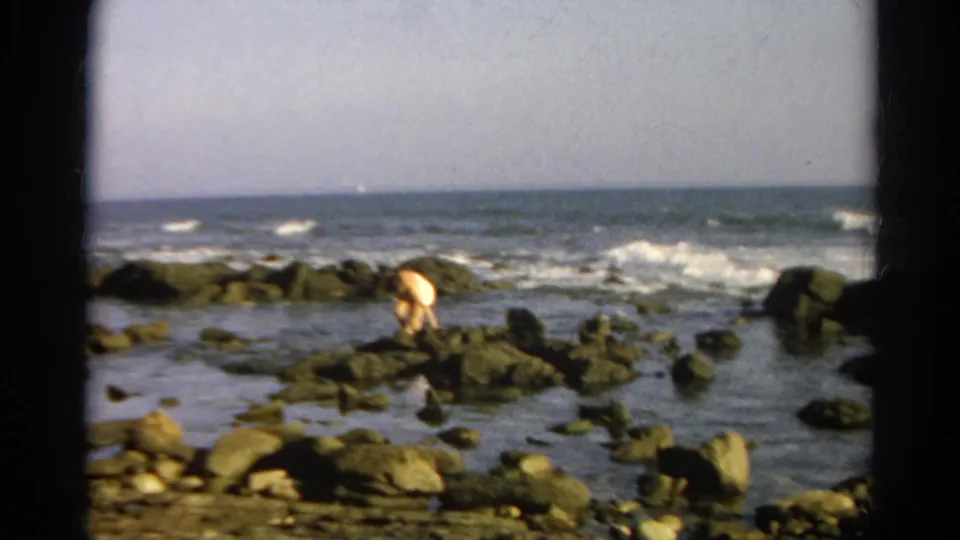 A soft 8mm frame shows the person leaning over dark coastal rocks, with shallow water and low surf in the background.