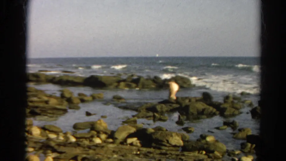 The final 1966 8mm frame keeps the person small in the scene, standing among rocks as the ocean breaks behind them.