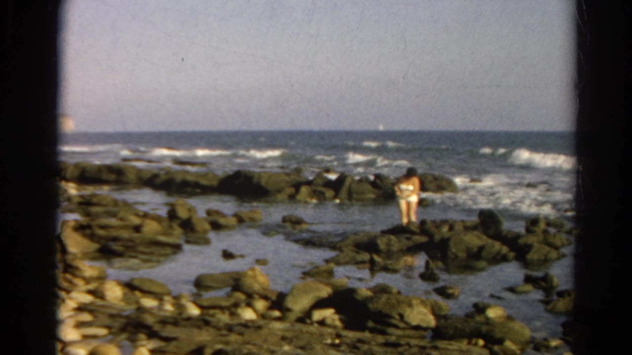 1966 8mm frame of a person in a light swimsuit standing on a rocky shoreline with waves behind them.