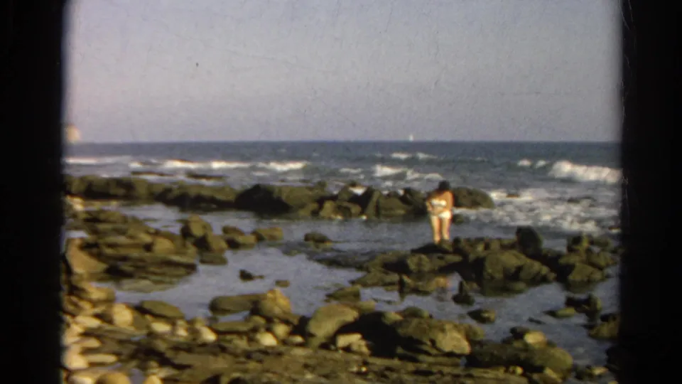 1966 8mm frame of a person in a light swimsuit standing on a rocky shoreline with waves behind them.