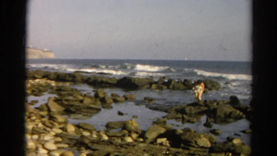 1966 8mm frame showing a distant person on coastal rocks, with surf, horizon, and dark film-frame borders visible.