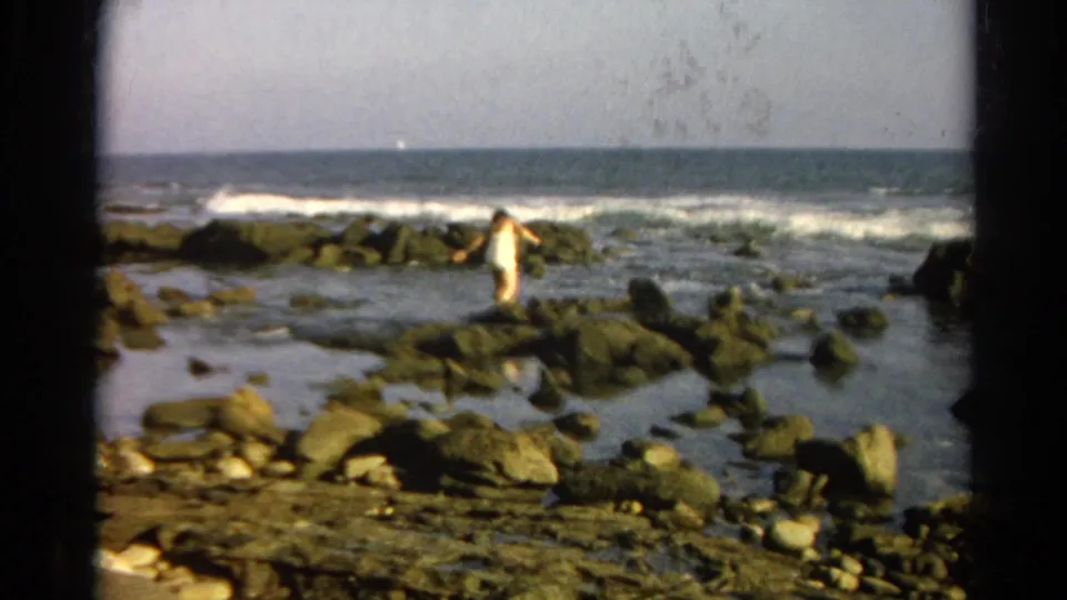 1966 8mm frame of a person navigating wet rocks along a shoreline with waves and tide pools behind them.