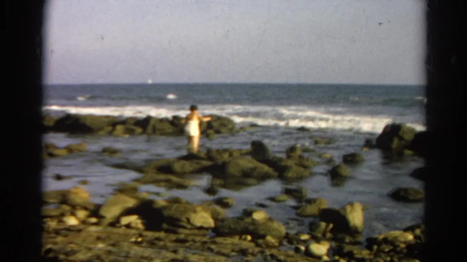 1966 8mm frame with a person centered on shoreline rocks, surf breaking behind them under a pale sky.