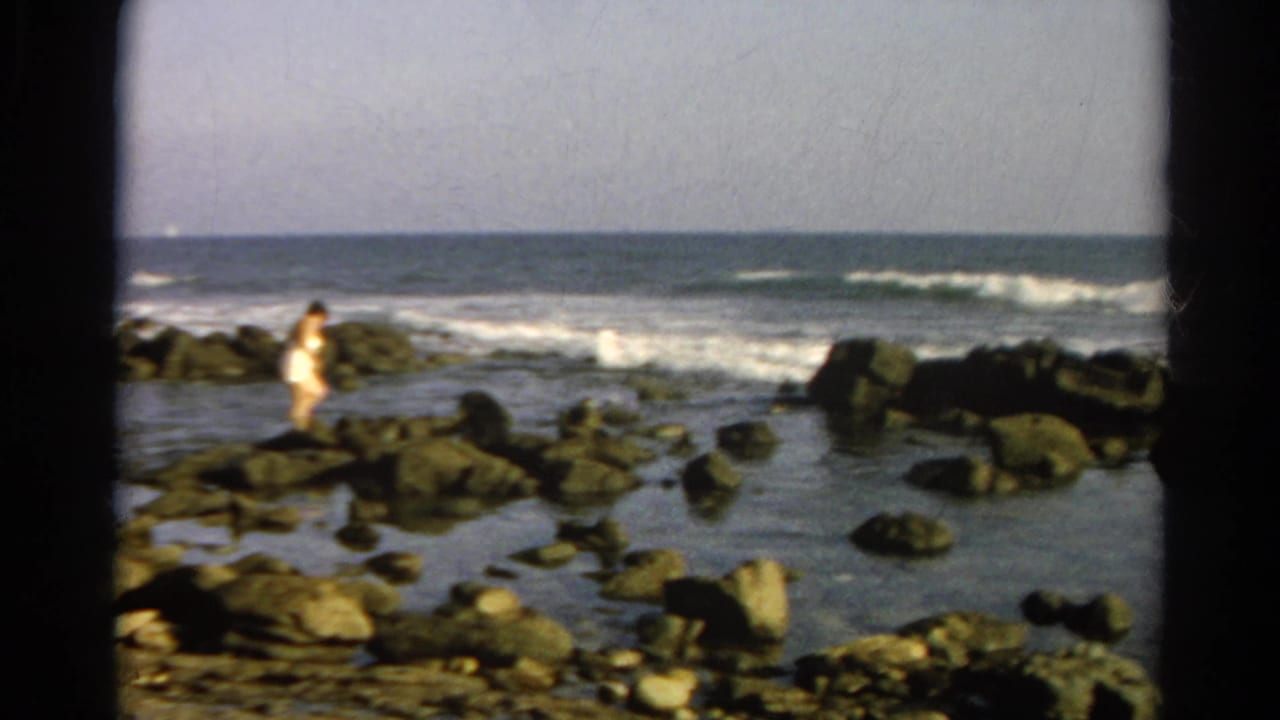 1966 8mm home-movie view of a rocky shoreline with tide pools, surf, pale sky, and a distant person near the water.