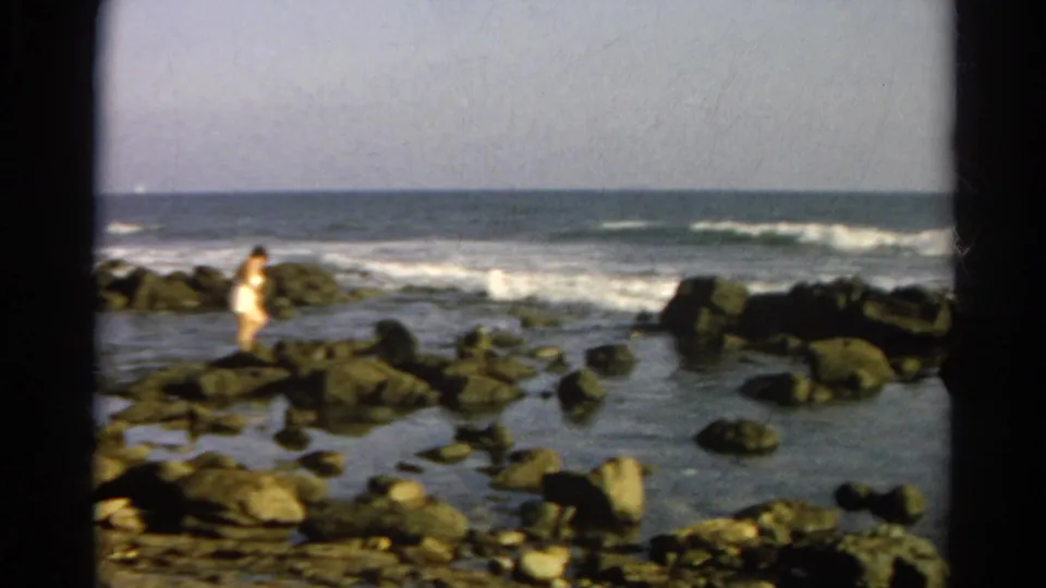 1966 8mm home-movie view of a rocky shoreline with tide pools, surf, pale sky, and a distant person near the water.