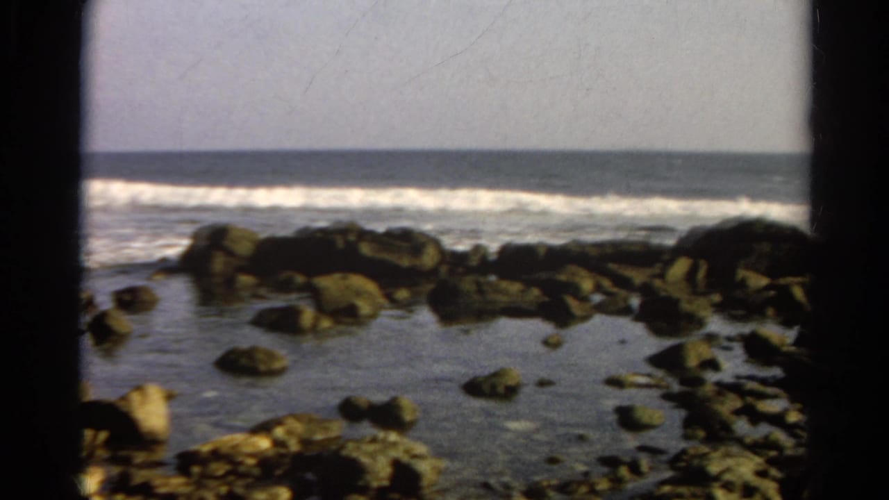 1966 8mm home-movie frame showing dark shoreline rocks, shallow tide pools, ocean surf, and a flat horizon.