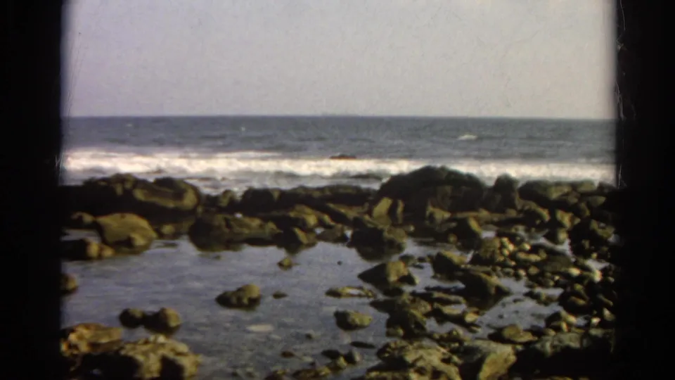1966 8mm home-movie frame of tide pools and scattered rocks along a shoreline with white surf beyond.