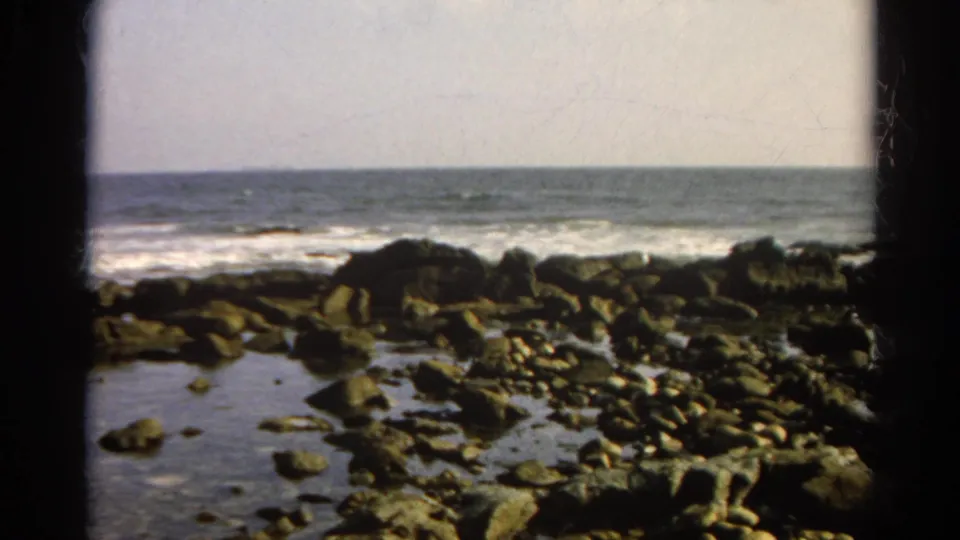 1966 8mm home-movie frame showing rocky tide pools and surf along a quiet shoreline under a pale sky.