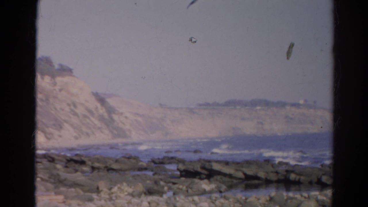 Rocky beach and surf below pale coastal cliffs, framed by dark vertical borders.