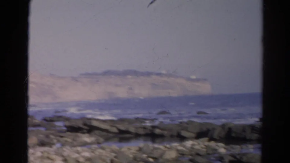 1966 8mm frame looking across rocks and surf toward a distant pale cliff line on the California coast.
