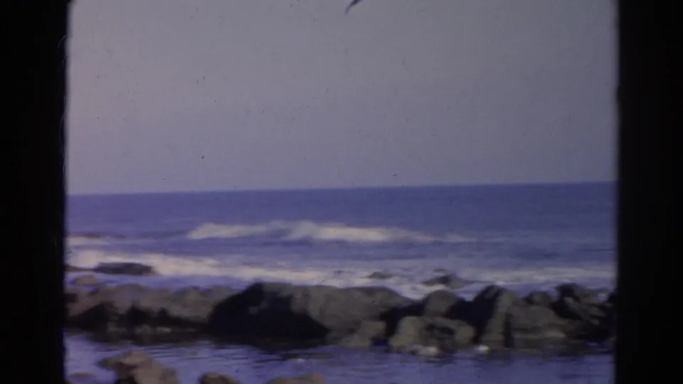 1966 8mm coastal frame showing open water, white surf, and dark shoreline rocks with soft focus and muted color.