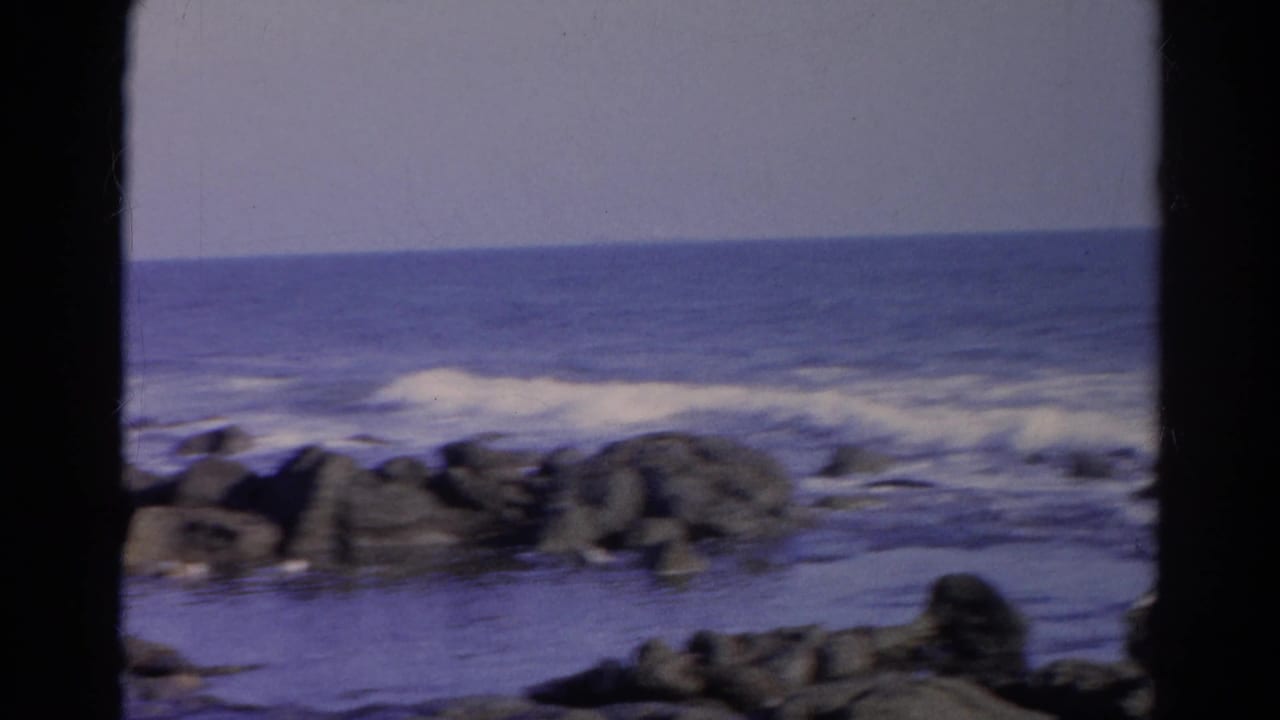 Small waves break behind dark rocks, framed by black 8mm film borders.