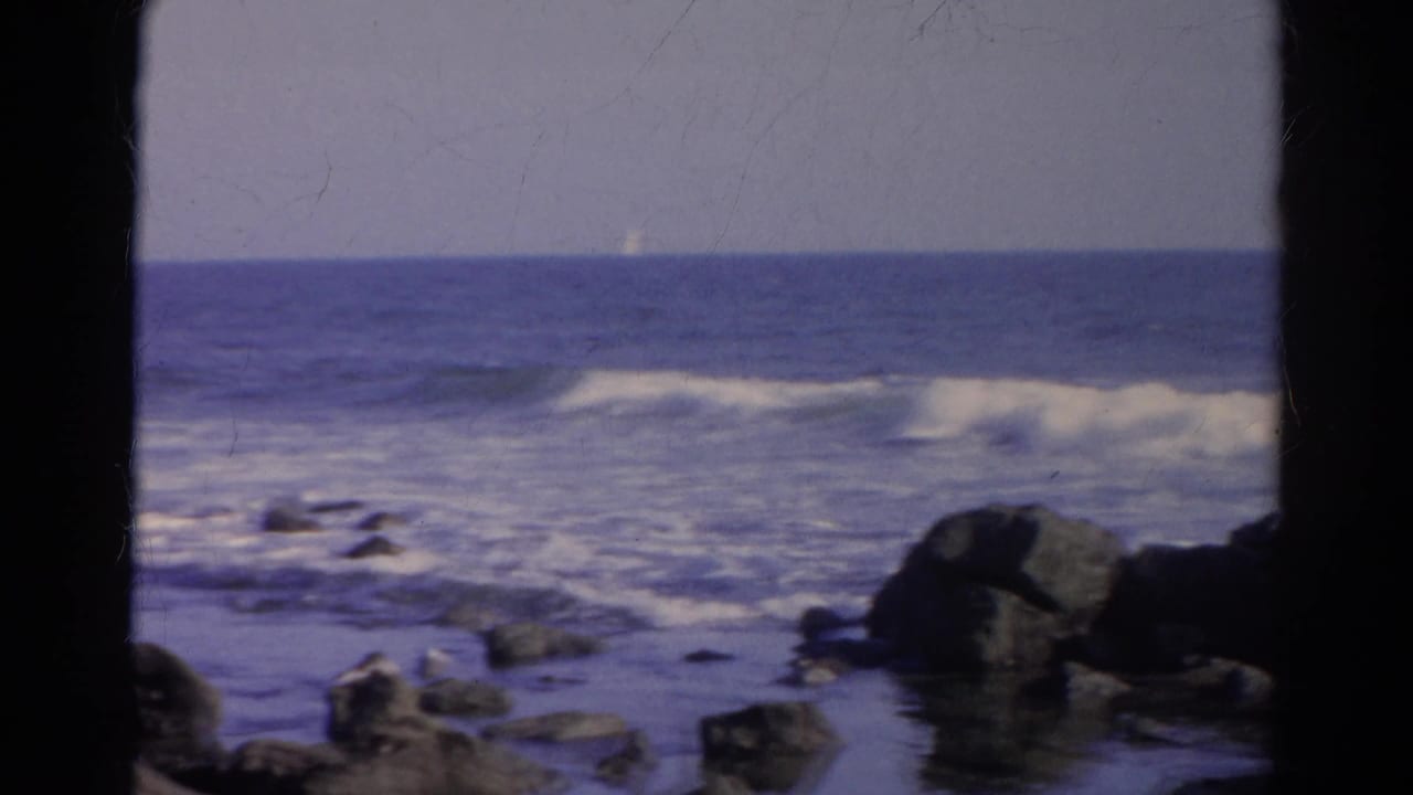 Rocky shoreline and low surf with a tiny sail-like shape near the horizon.