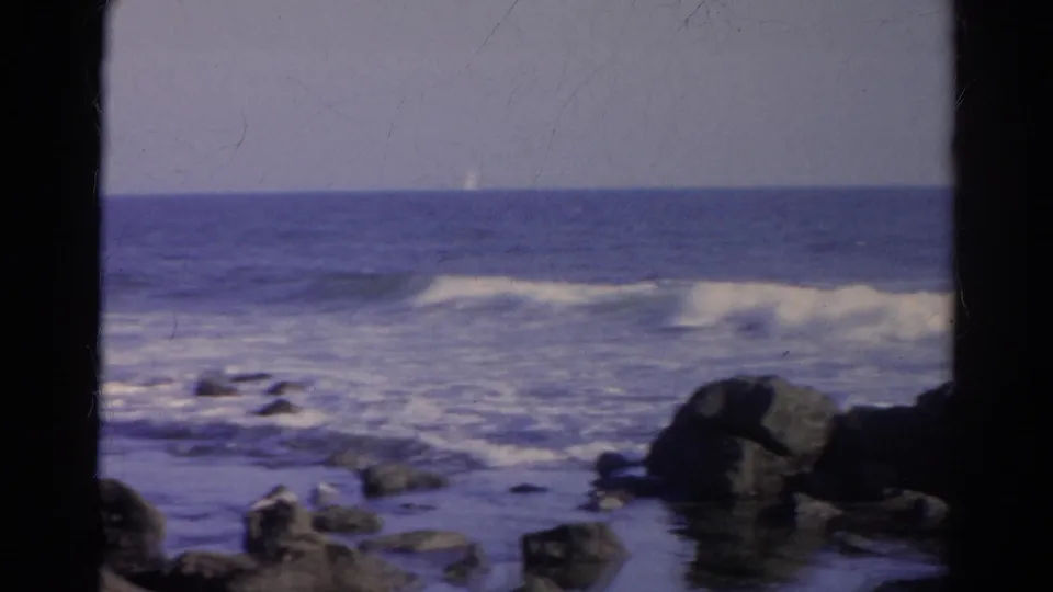 1966 8mm shoreline view identified as Silver Lake, California, with rocks, low waves, and a small distant sail-like shape on the horizon.