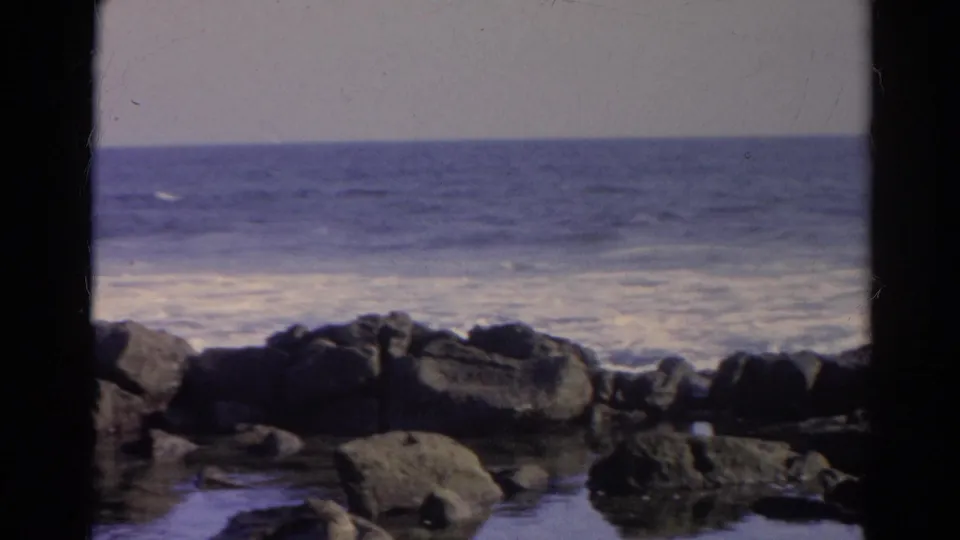 1966 8mm shoreline frame identified as Silver Lake, California, with shallow water, dark rocks, distant surf, and a muted blue-purple cast.