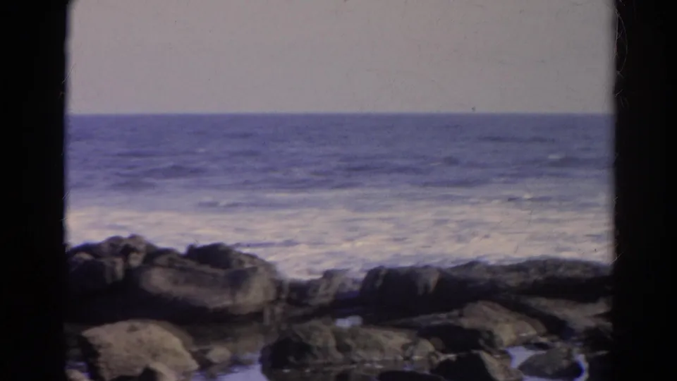1966 8mm home-movie frame showing waves rolling toward dark shoreline rocks under a soft horizon.