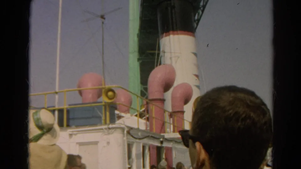1966 8mm home-movie view of a docked liner's funnels and railings with spectators partly blocking the foreground.