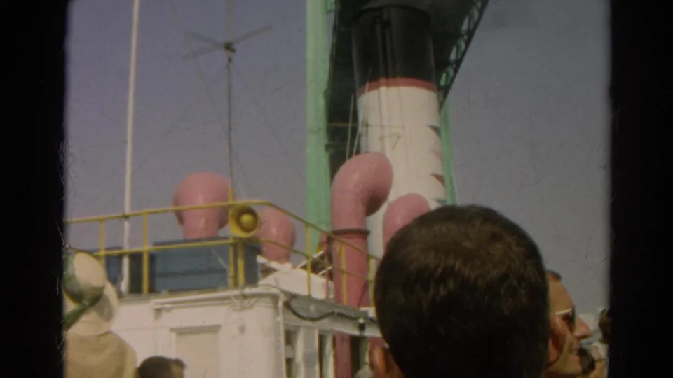 1966 8mm frame showing spectators beside a docked liner, with pink funnels, railings, and a smokestack above them.