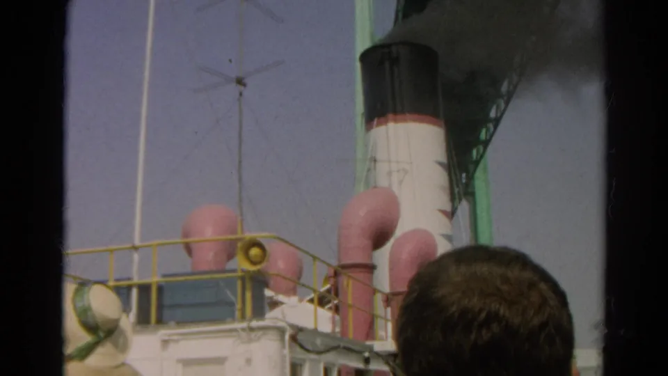 A handheld 8mm view shows the ship's deck railing, pink vents, smokestack smoke, and a spectator's head along the lower frame.