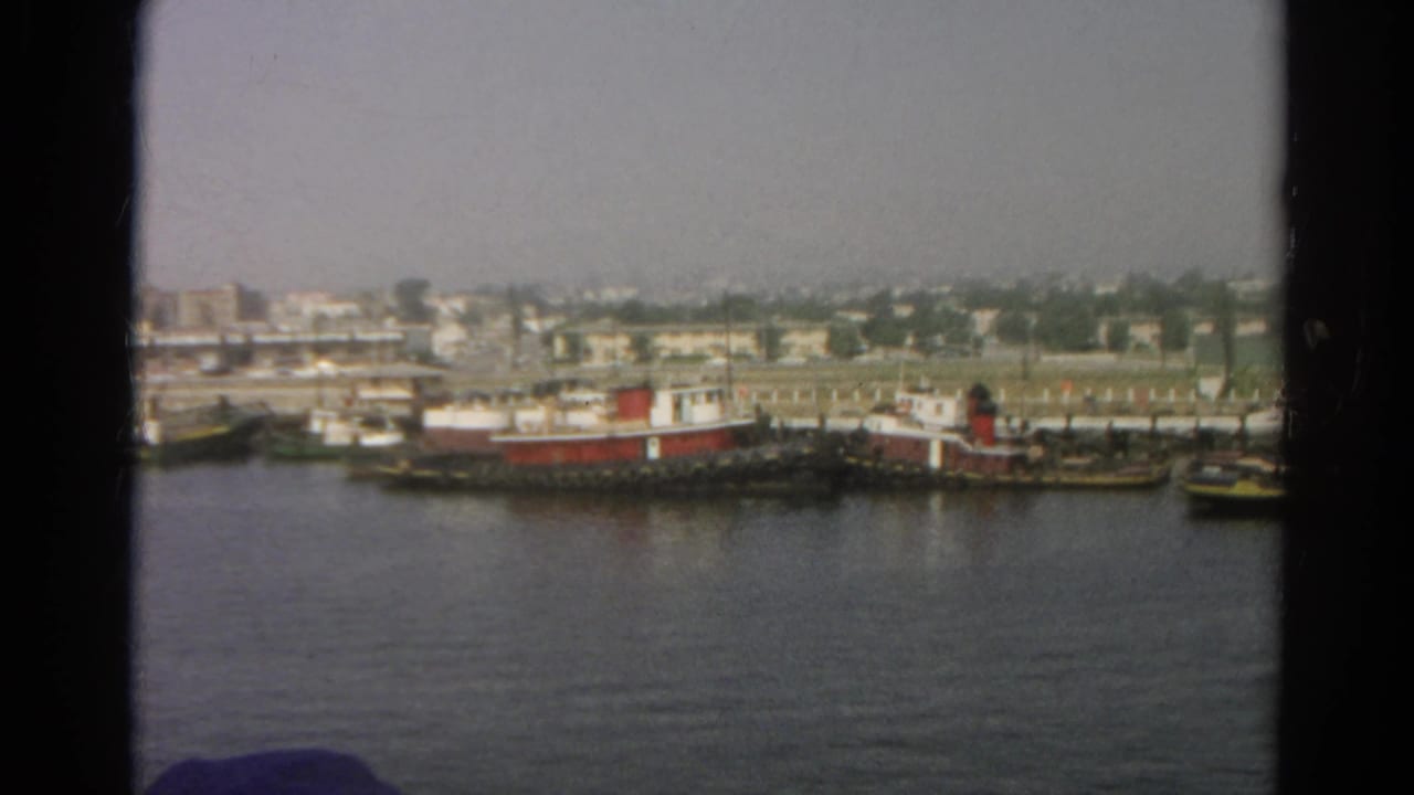 1966 8mm boat-view frame looking across water toward docked tugboat-like vessels, low buildings, and a hazy shoreline.