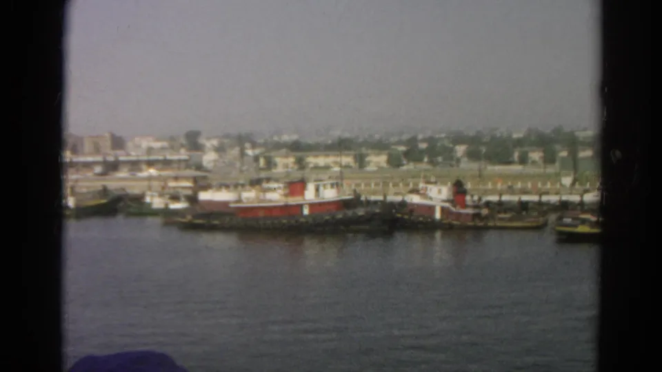 1966 8mm boat-view frame looking across water toward docked tugboat-like vessels, low buildings, and a hazy shoreline.