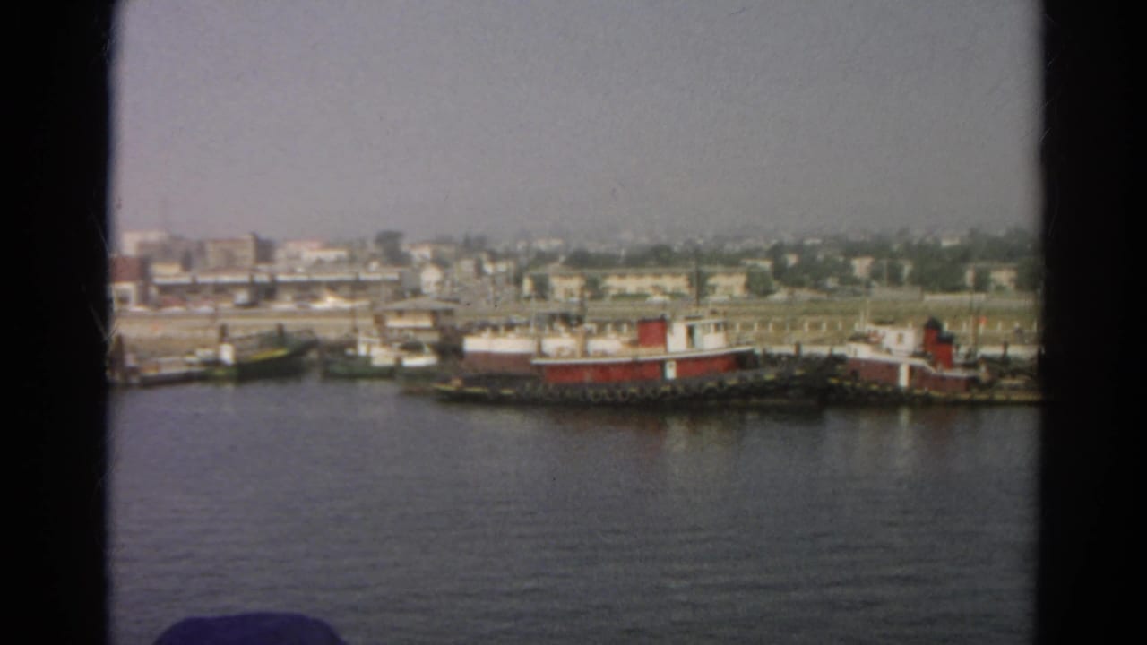 1966 8mm frame from a boat showing moored vessels, shoreline buildings, and a hazy California waterfront view.
