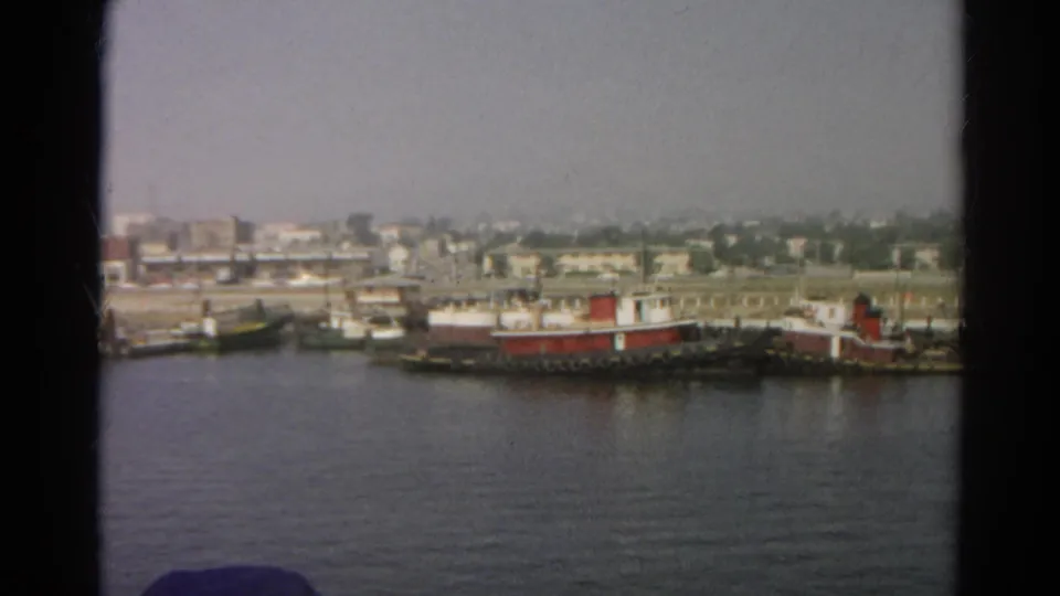 1966 8mm frame from a boat showing moored vessels, shoreline buildings, and a hazy California waterfront view.