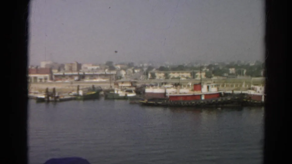 1966 8mm boat-view frame with water, docked boats, low harborfront buildings, and soft faded color.