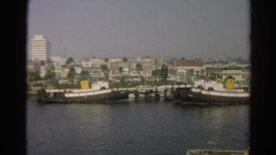 1966 8mm frame showing tugboat-like vessels near the shoreline, with palm trees, low buildings, and a taller white building behind.